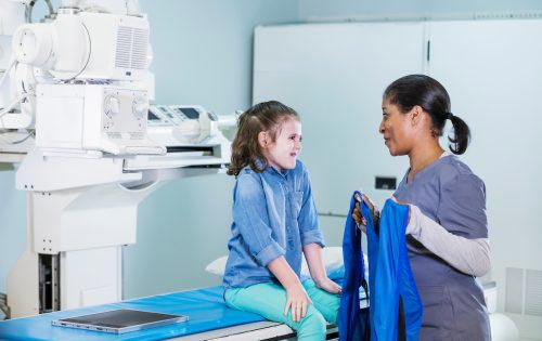 A medical professional in scrubs interacts with a young girl who is sitting on a medical table in a radiology or medical imaging room. The professional is holding a protective lead apron, preparing the girl for an X-ray or similar procedure.