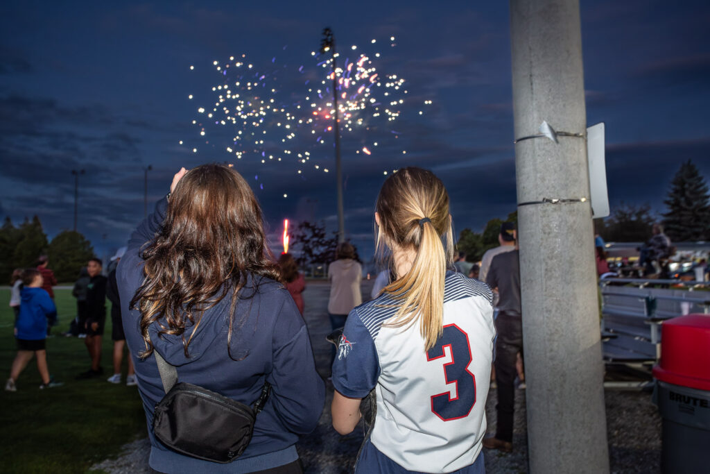 Two students standing side by side, watching a vibrant fireworks display light up the night sky during a campus event. The scene captures the excitement and celebration, with one student wearing a jersey marked with the number 3, adding a sense of school spirit to the moment.