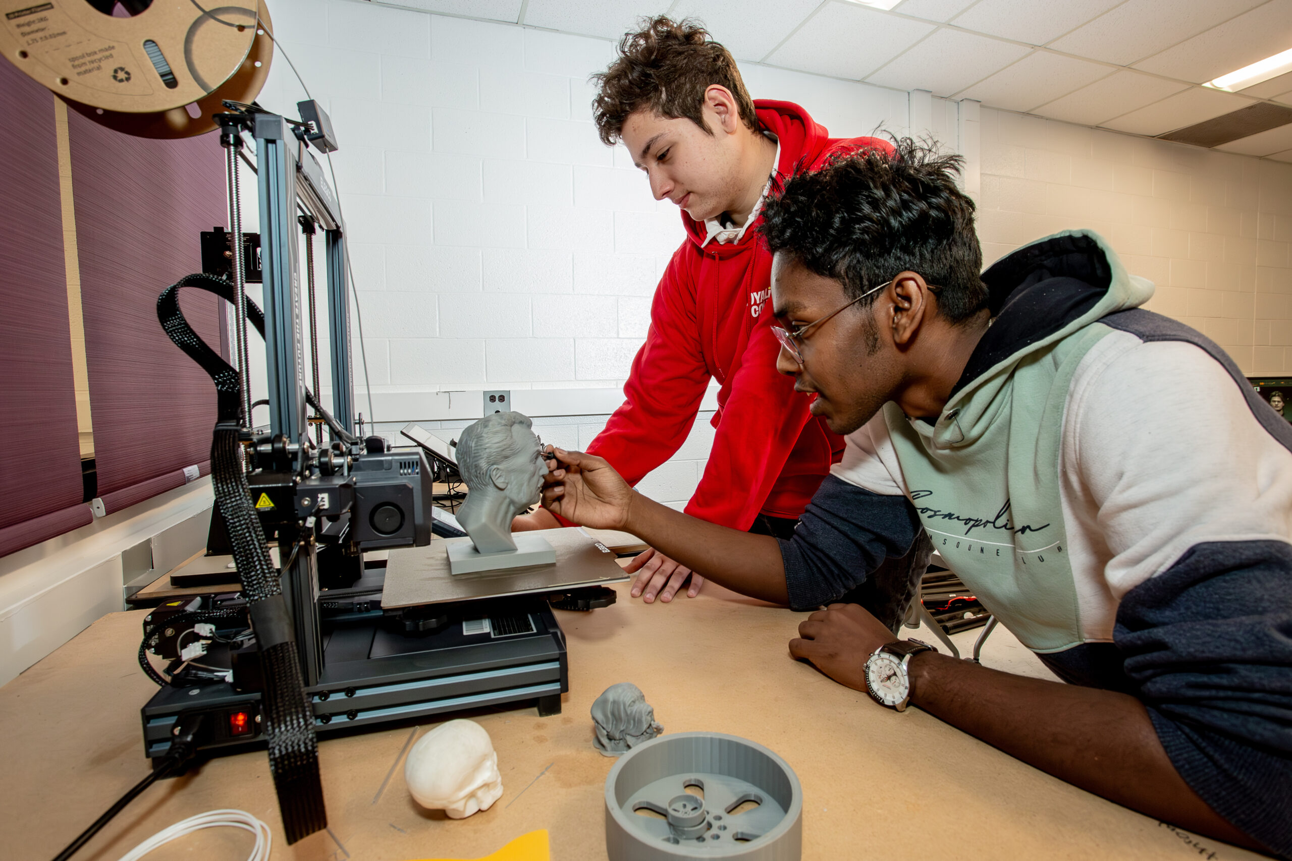 Two students are deeply engaged in a 3D printing project in a lab at Loyalist College. One student is carefully examining and adding details to a small, intricately printed bust, while the other monitors the progress of the 3D printer. Various 3D printed objects, including a small skull and a mechanical part, are scattered on the workbench, showcasing the practical and creative aspects of their work. The scene captures the hands-on, collaborative learning environment that encourages innovation and exploration in technology at Loyalist College.