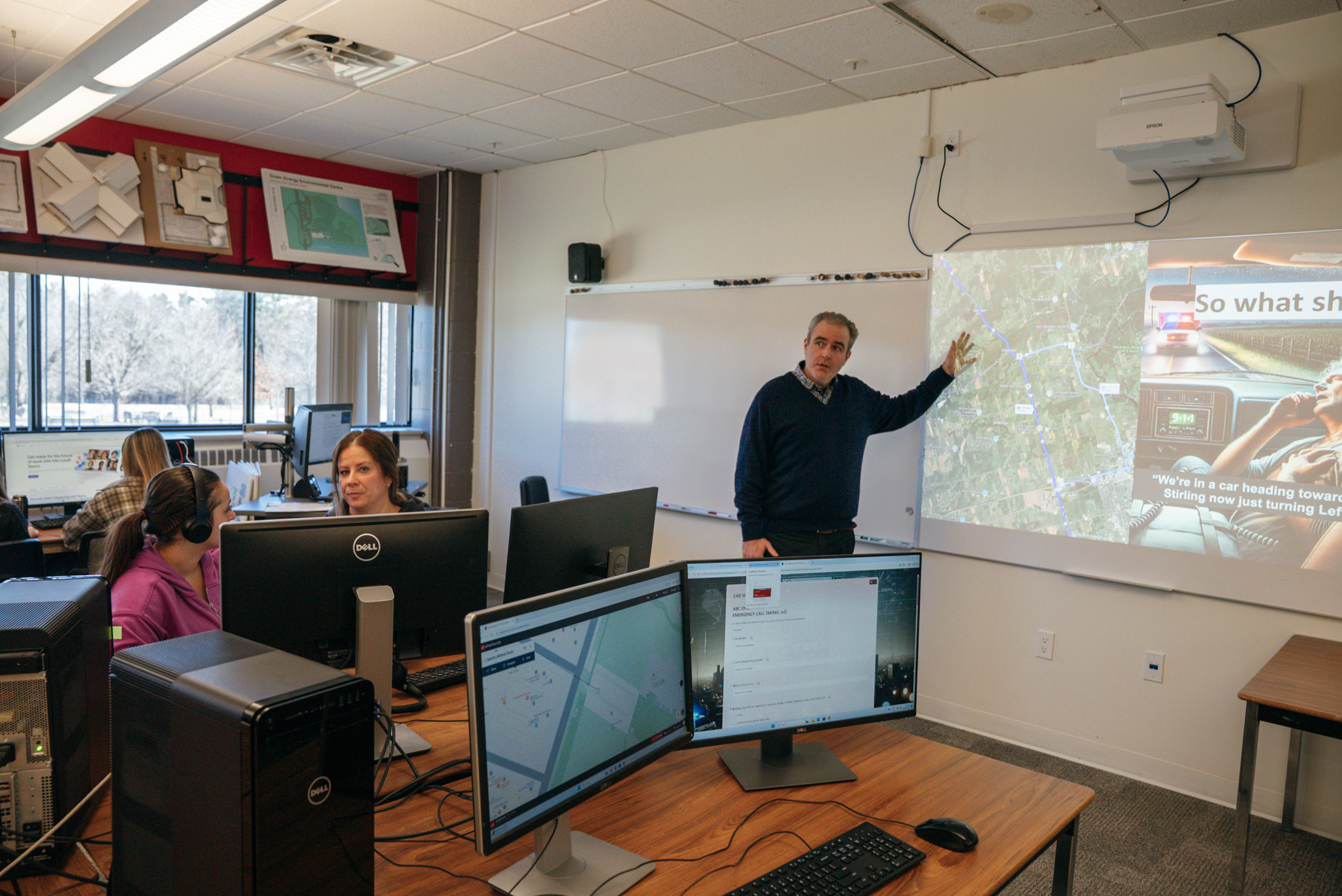A male teacher is standing at the front of the room, pointing at images being projected onto the board behind him. On the board is images of a map, as well as an image and text cut off on the right side. Students sit at computers around the room.