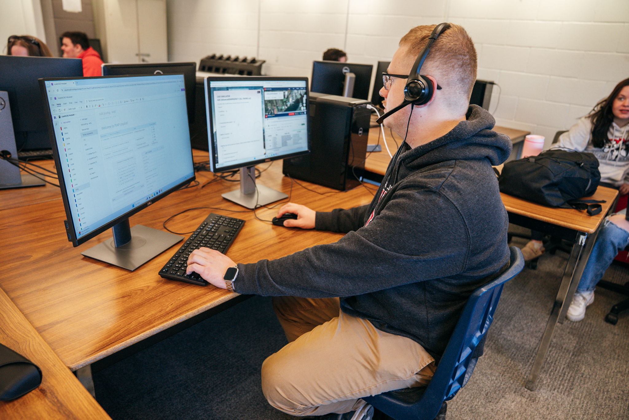 A man sits at a desk in a classroom, with two monitors in front of him. He has one hand on the keyboard, the other holding the mouse. He's wearing a headset as he prepares to take a mock emergency call.