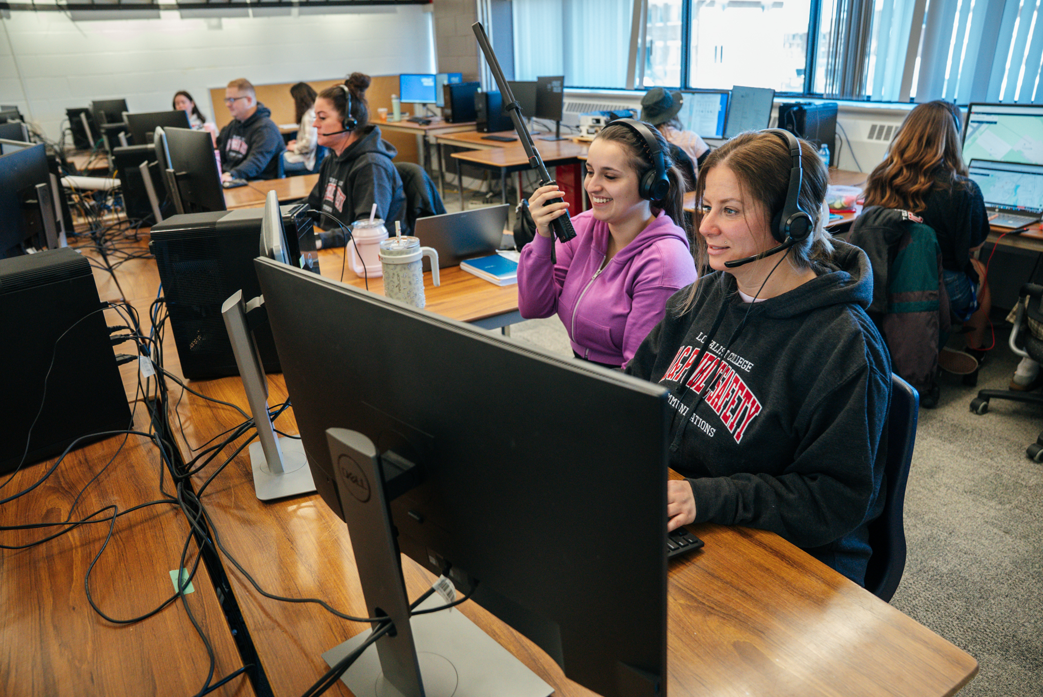 Two girls sit at a desk in a classroom in front of a computer. The one girl is using the keyboard and mouse, while wearing a headset. The other girl is slightly off to the side with a large walkie-talkie radio in her hands, raised to her mouth. Other students can be seen in the background, also sitting at computers with headsets.