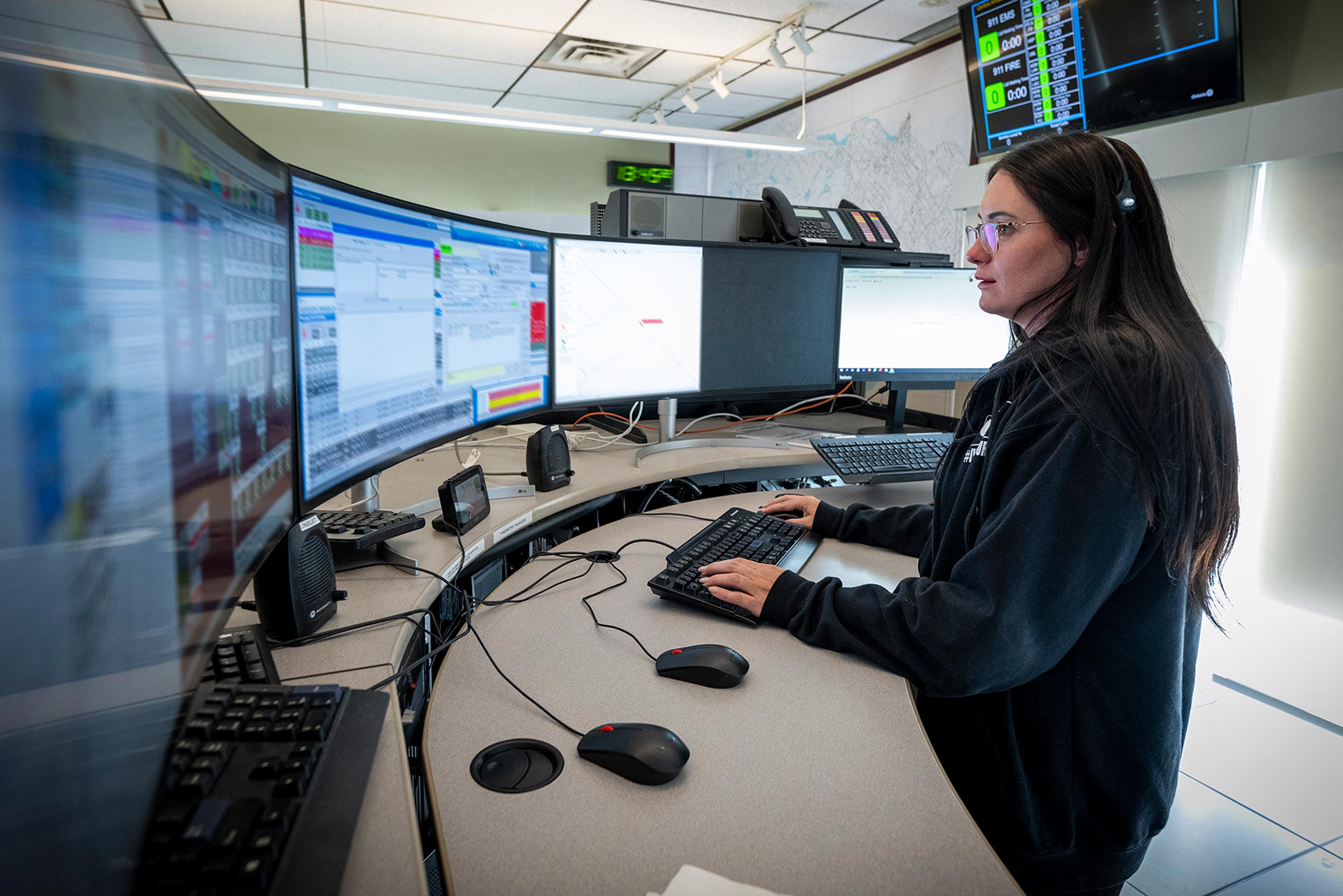 A female dispatcher wearing a headset is standing at a command centre with multiple large monitors displaying emergency response data. She is focused on her screen, managing various communications and coordinating emergency responses.