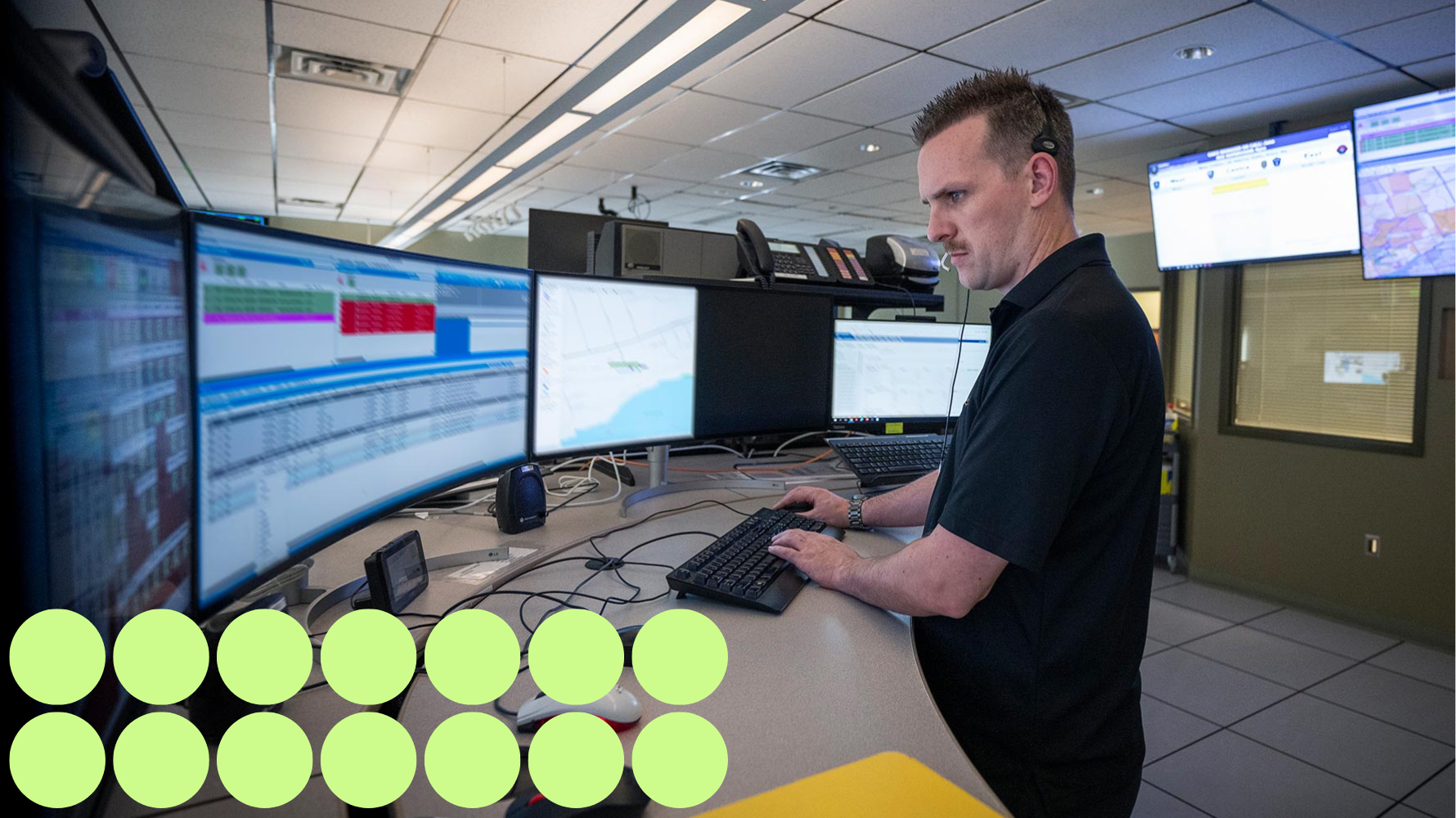 A 911 dispatcher at work in a control room, focused on multiple large computer monitors displaying maps and call information. The dispatcher wears a headset and types on a keyboard, managing emergency communications.