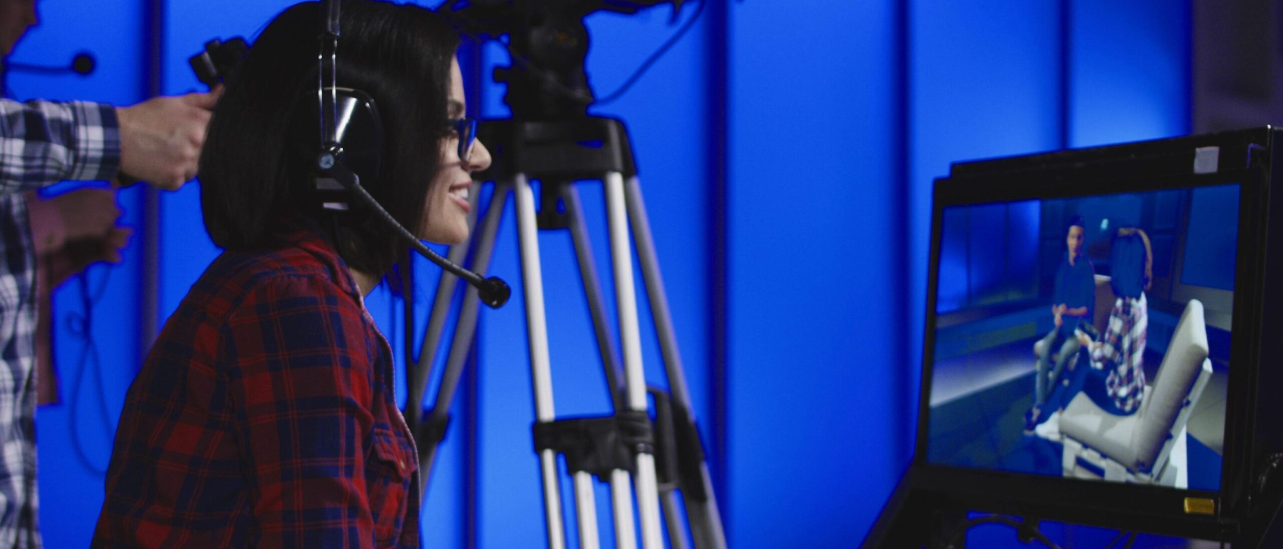 A studennt is wearing a headset smiling and looking to the right of the image. Behind the student is another student operating a video camera. On the right of the image is a display showing a live film production of people talking.