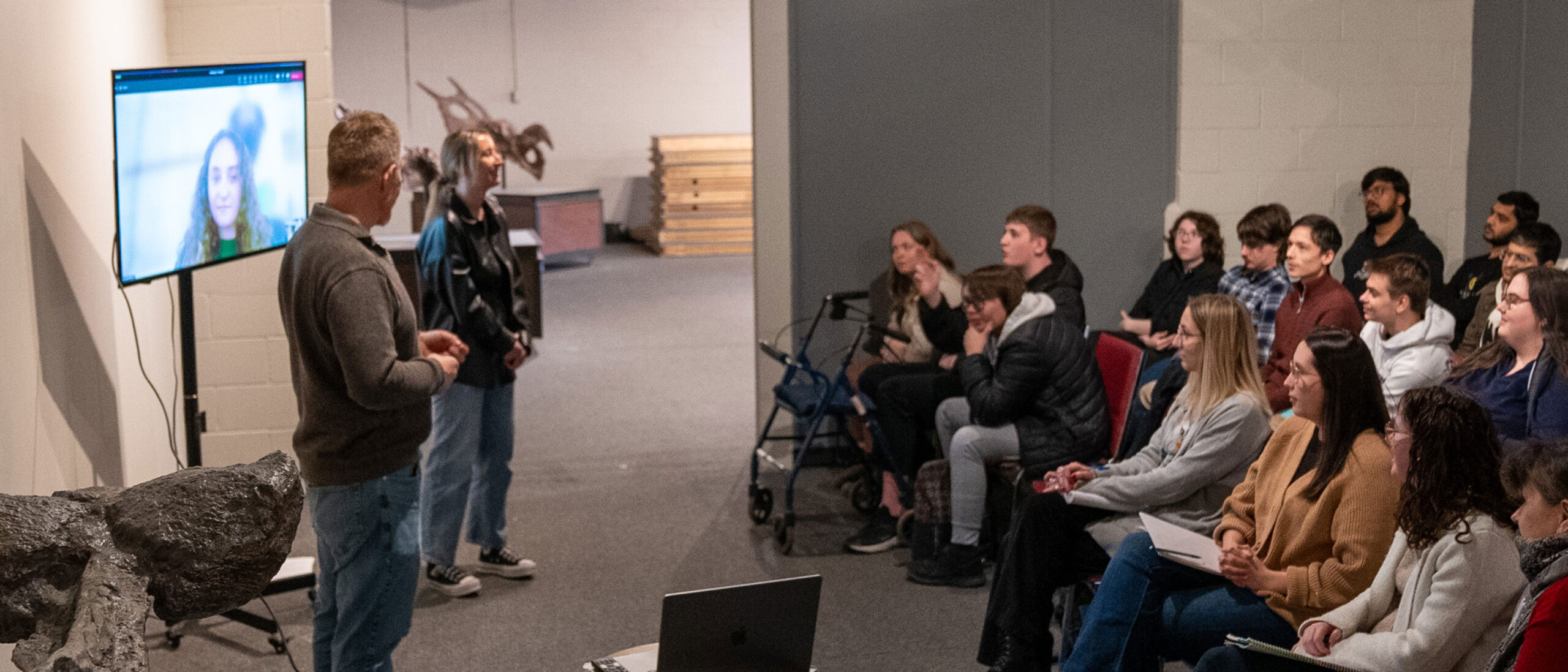 Two people are presenting to a group of students in a lecture hall or classroom setting.