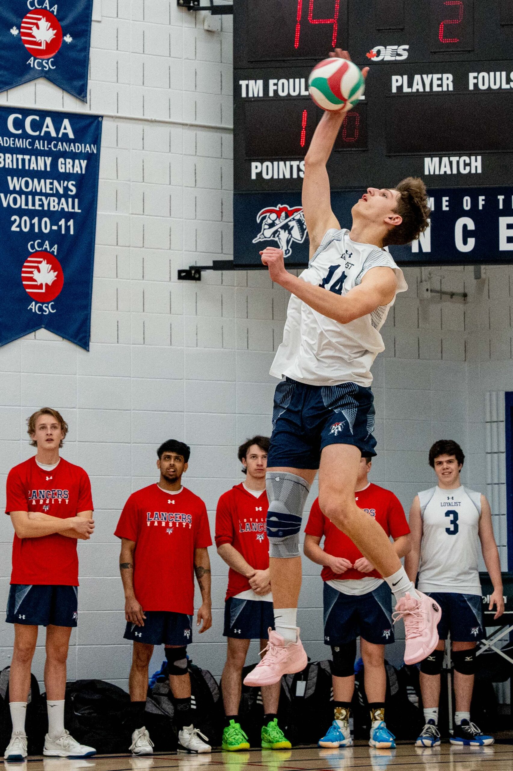 A student jumps into the air to serve a volleyball, while other students watch from the sidelines. The scoreboard and championship banners can be seen in the background.