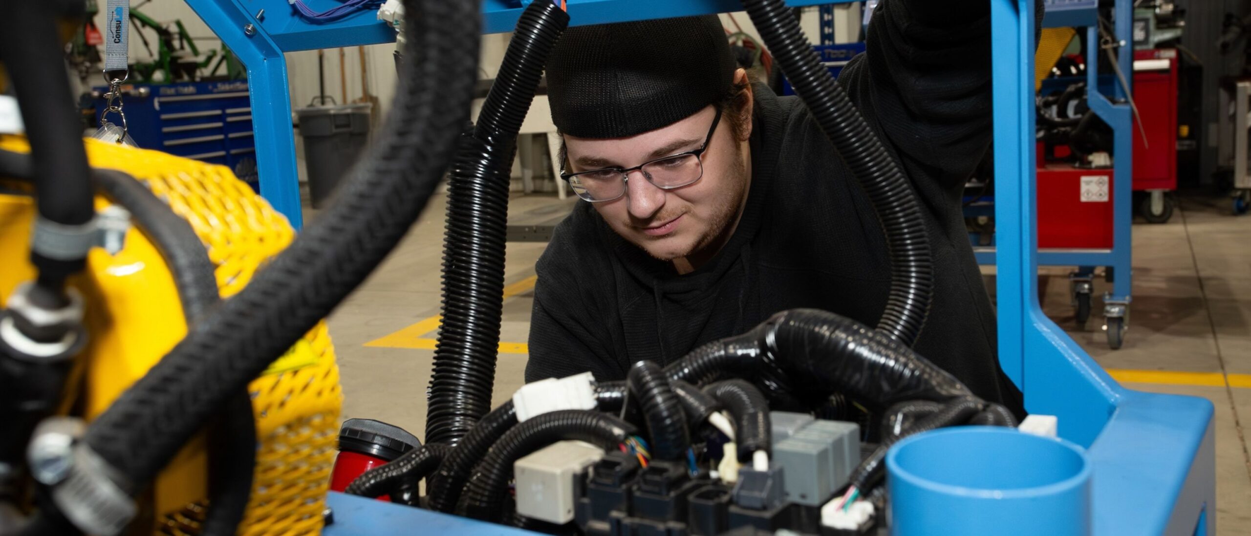 A student crouches down in front of a work trolley, looking at automotive components within a service shop.