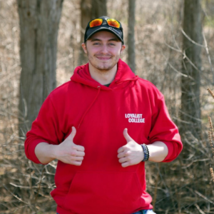 A headshot of Bradley Dall-Smith, a CYC student, in a red Loyalist sweater. He's holding two thumbs up and in the background is a wooded forest.