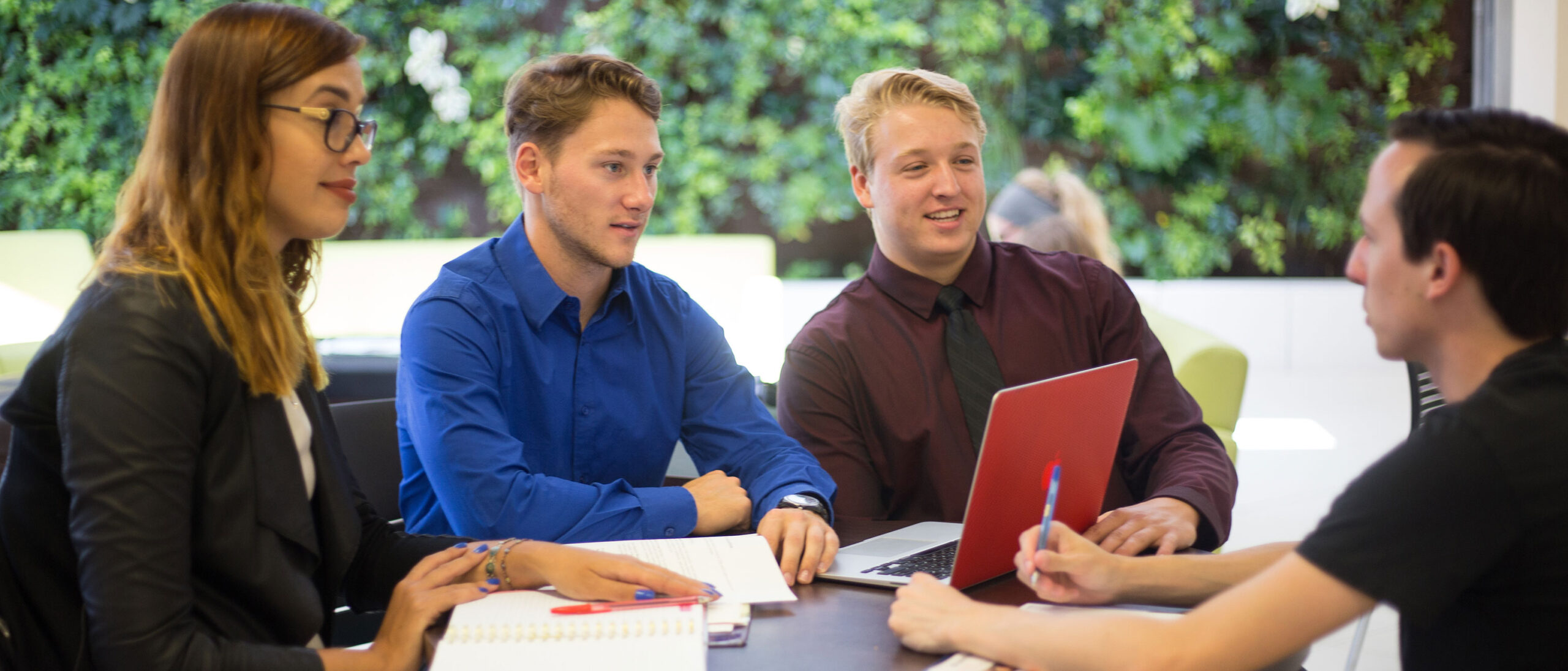 A group of four students are sitting around a table wearing business professional clothing and engaging in conversation with one another. There is a bright green botanical wall display behind the students.