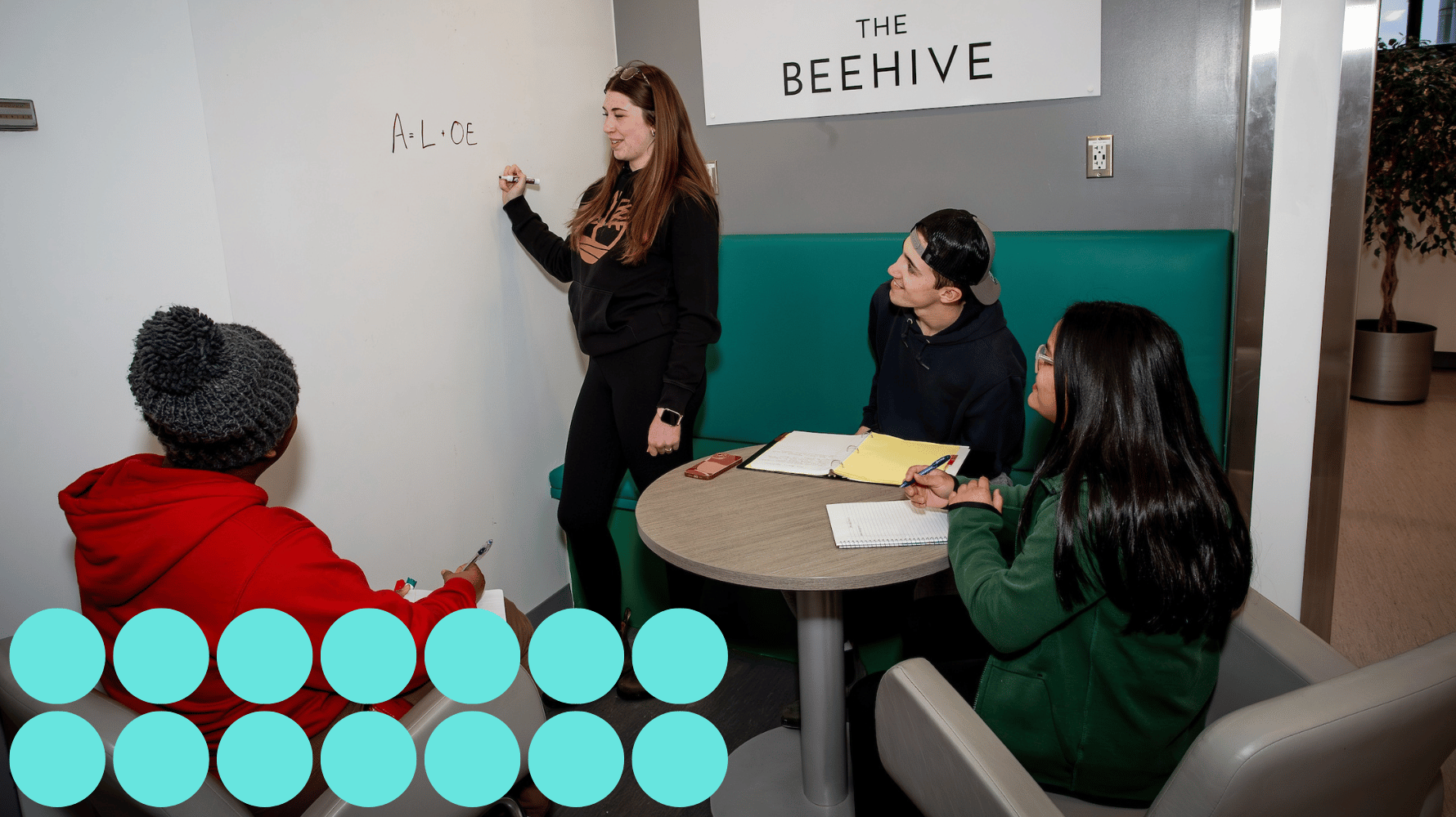 Three students are sitting down while looking at a fourth student standing. The standing student is writing on a whiteboard. Fourteen blue graphic circles are in the lower lefthand corner of the image.