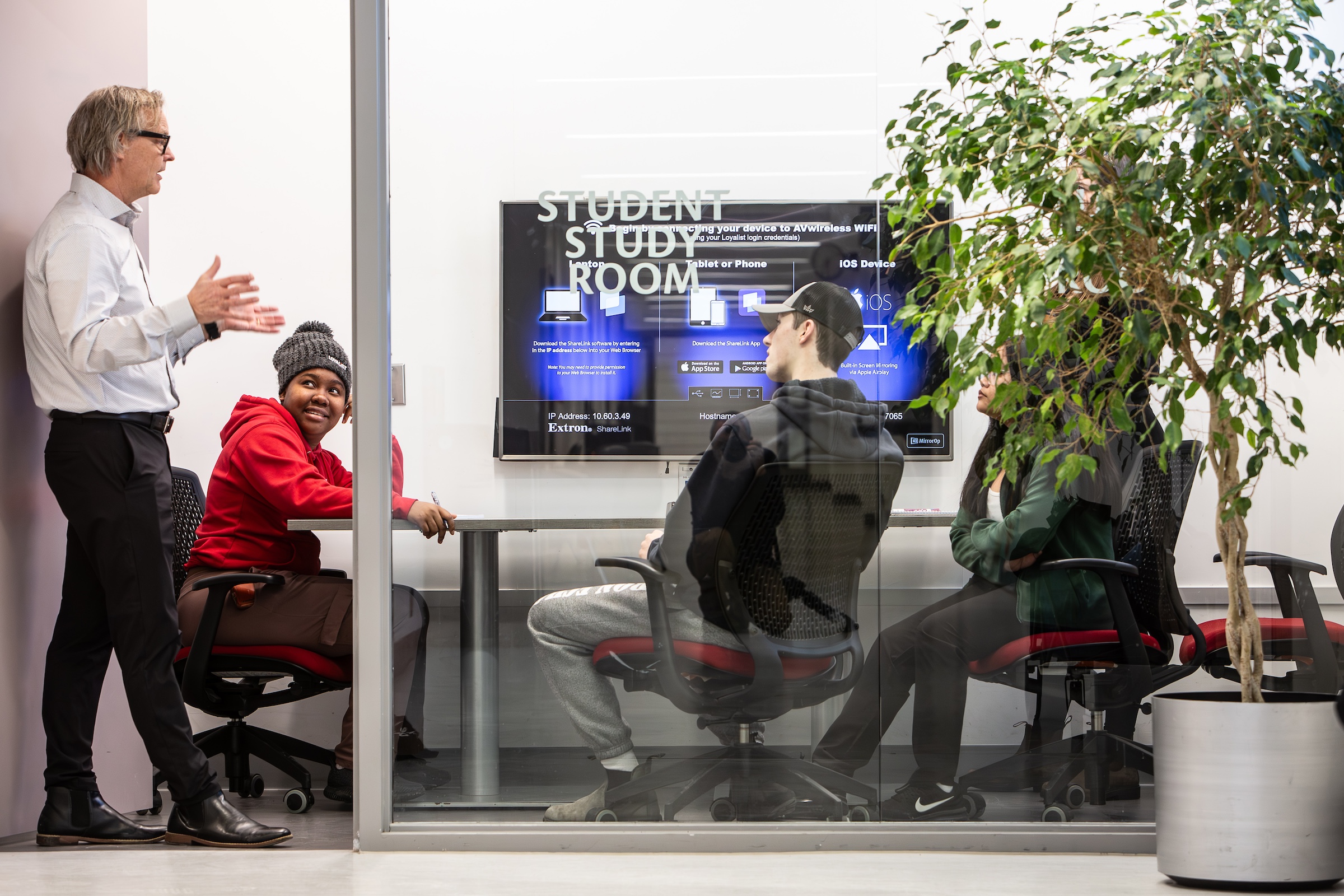 A group of students and faculty member are inside the student study room. The students are sitting at a desk with a television screen in front of them. The faculty member is standing by the doorway speaking to the group.