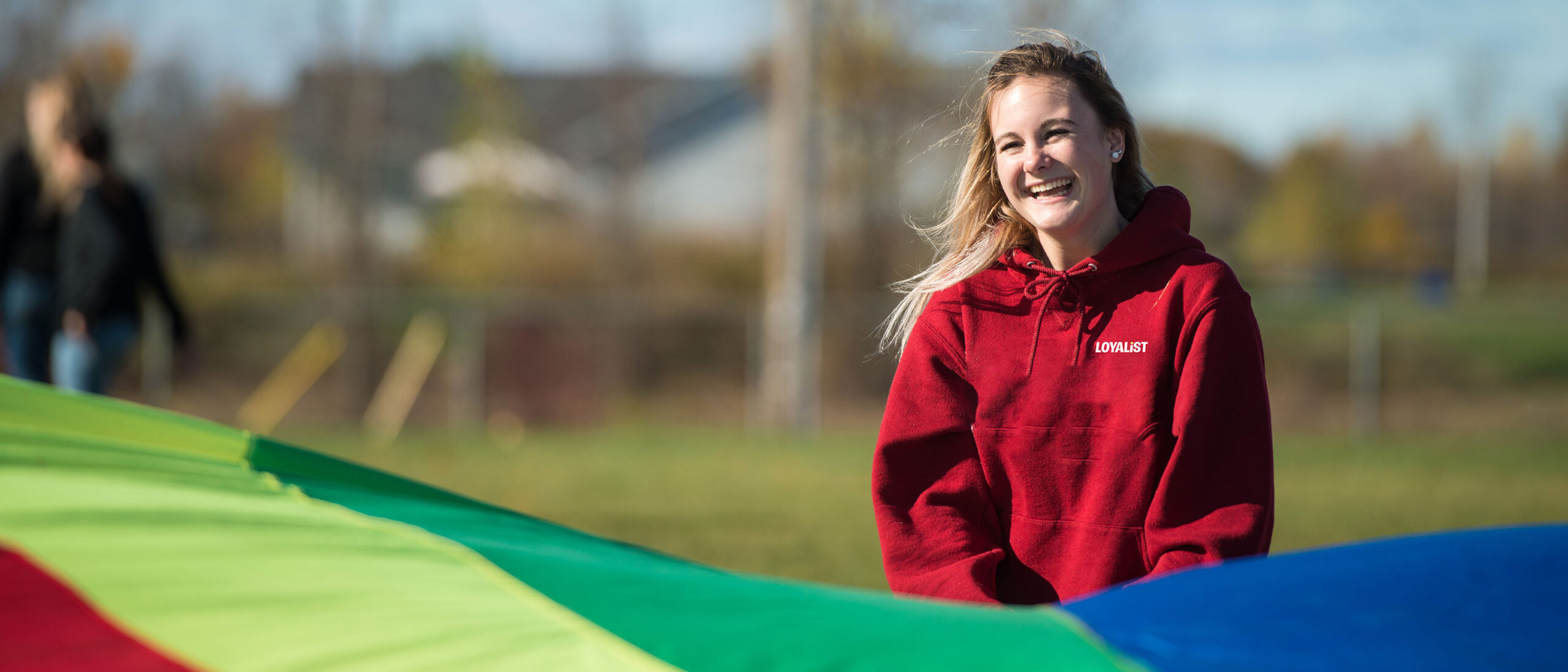 A happy student in a red Loyalist sweatshirt is standing outside holding a large multicoloured tarp.