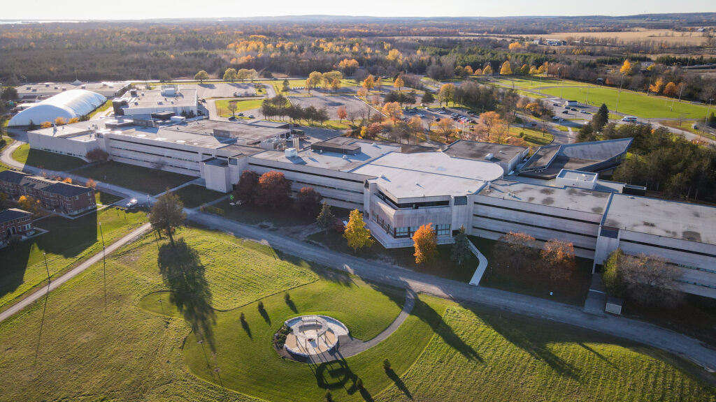 An aerial view of the Loyalist College Belleville campus. A large white building sprawls over green grass with trees in the background.