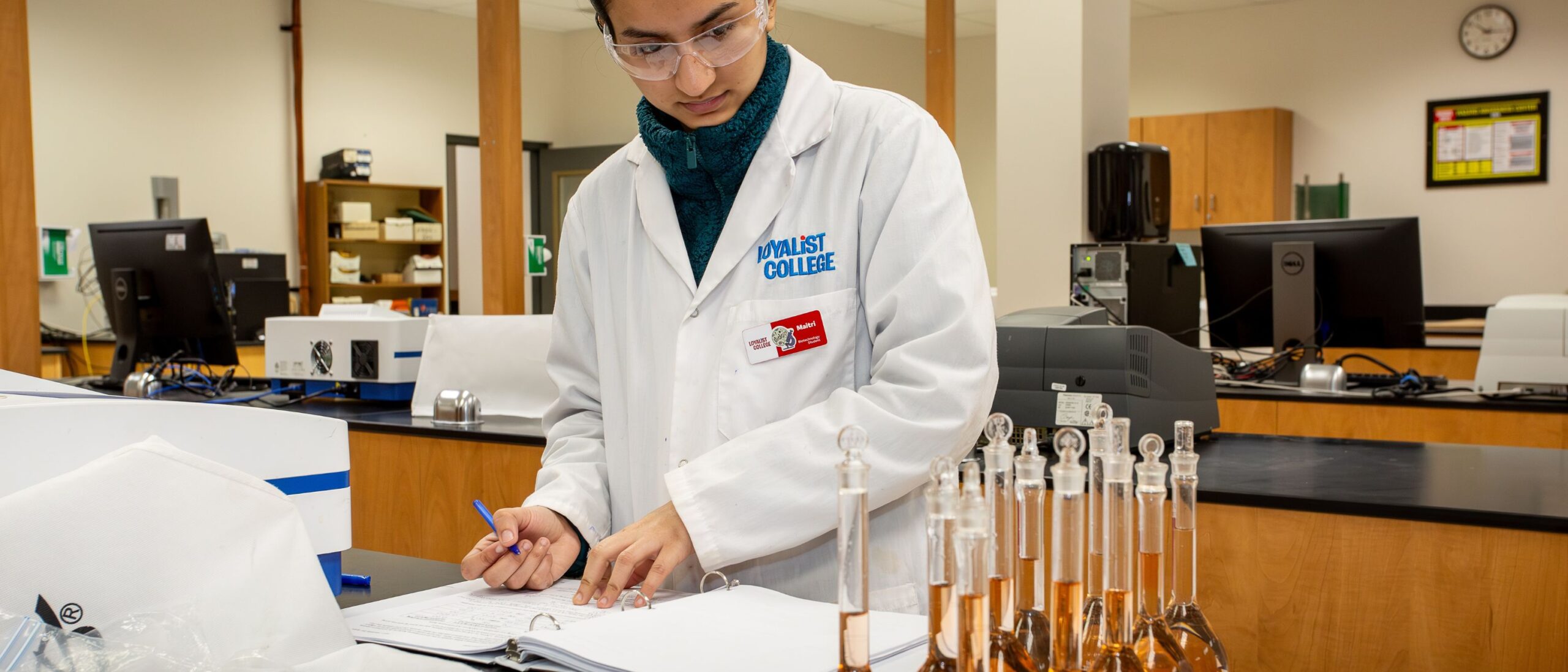 A chemical engineering technician is wearing a pair of goggles and a Loyalist College white lab coat. The student is looking at test tubes on their left with their hands over a binder full of paper. The student's right hand is holding a blue pen.