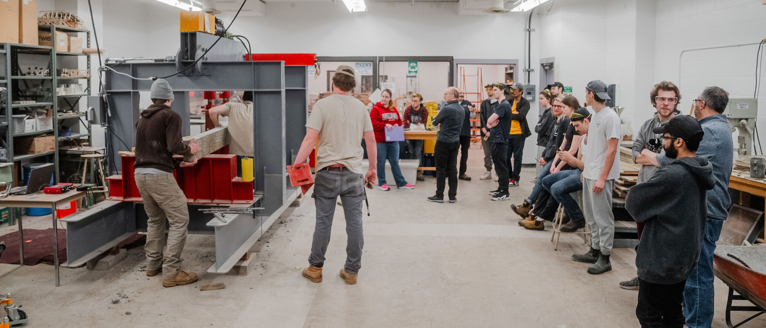 A group of civil engineering technician students are located in a classroom setting. Some students are actively working with wood and machinery, focusing on their tasks, while the rest of the group observes, attentively watching the process