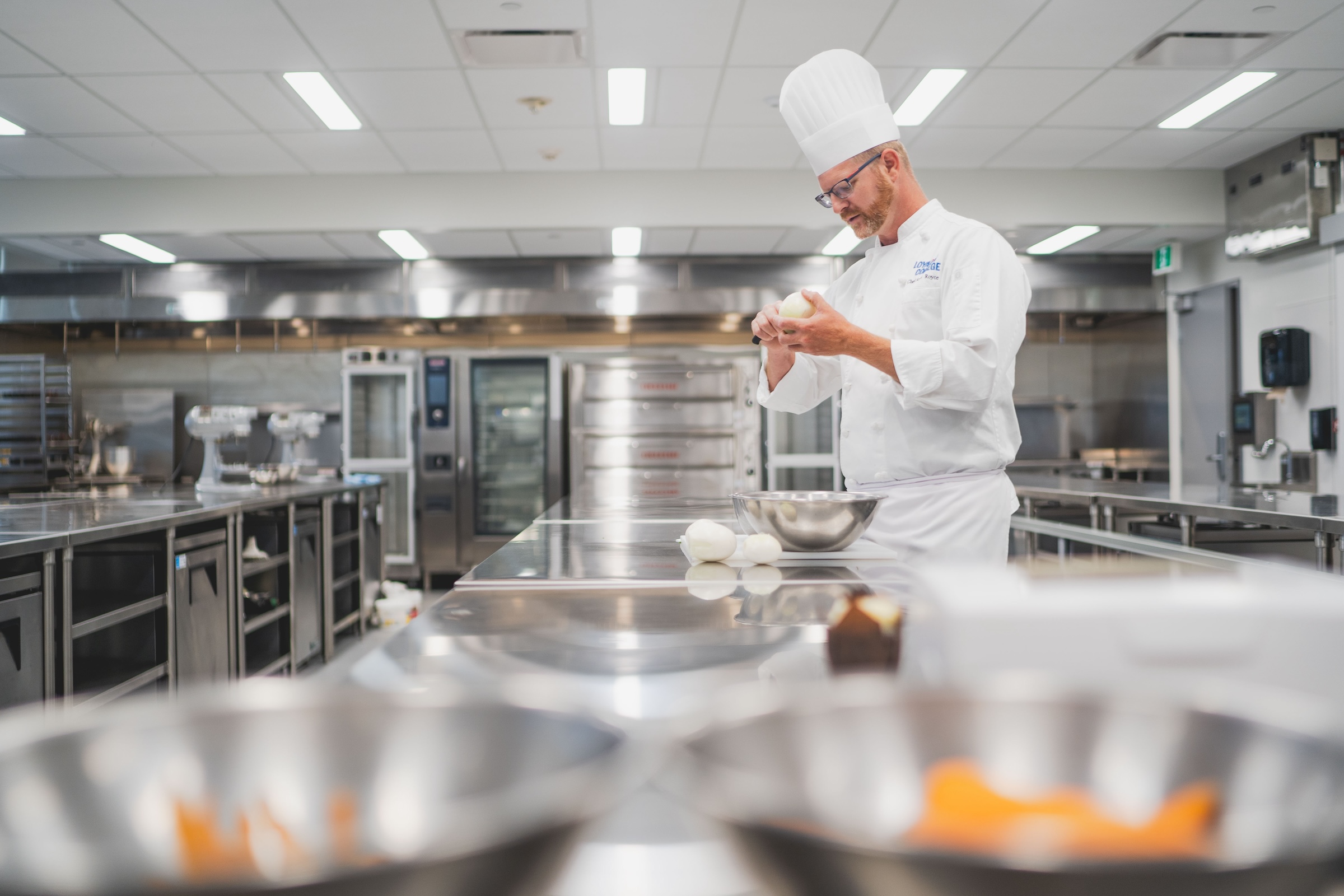 A chef in a chef's hat and jacket works over a large bowl on a counter in the culinary lab.