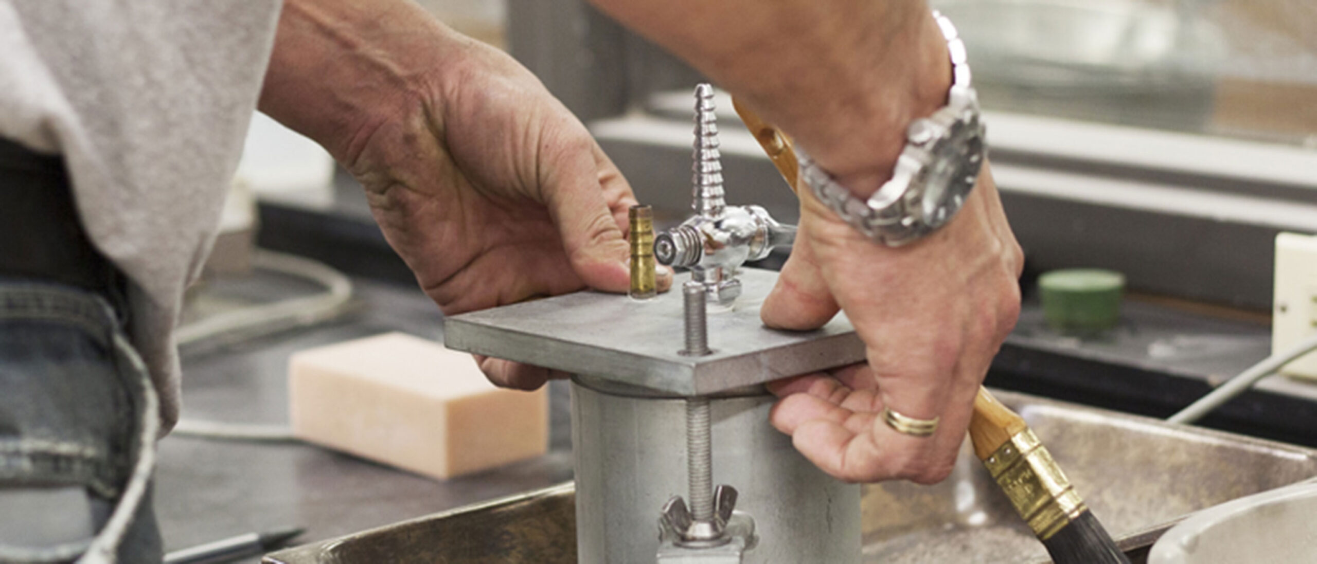 Close-up of a Construction Engineering Technician's hands using a tool to degas paint. The person is wearing a ring on their right hand and a silver watch on their wrist, focusing on the precise task.