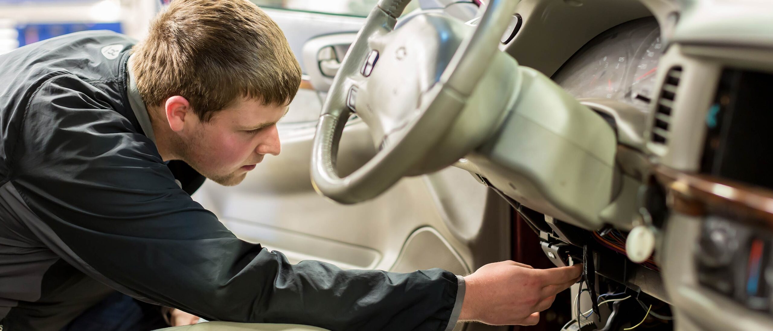 A customs border services student is wearing a long sleeved shirt, leaned forward and looking below a driver's steering wheel inside an automotive vehicle.