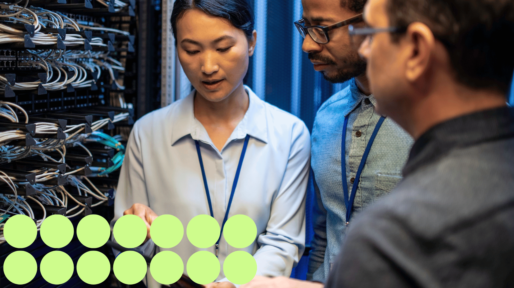 Three people wearing lanyards and business attire stand behind servers looking at a tablet. Green graphic circles are in the lower lefthand corner of the image.