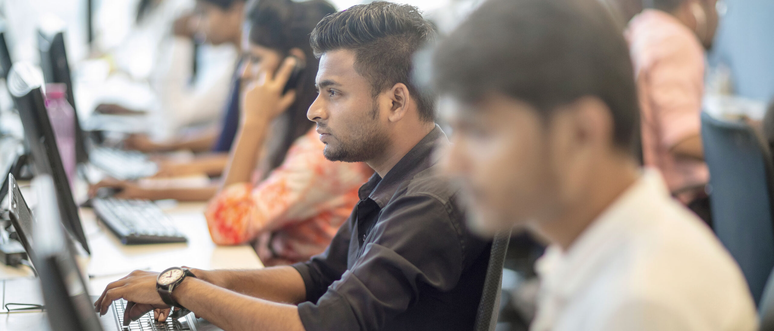 A group of Cyber Security students are working inside a computer lab sitting down in front of computers. The students are either on the phone, typing or reading off the computer.