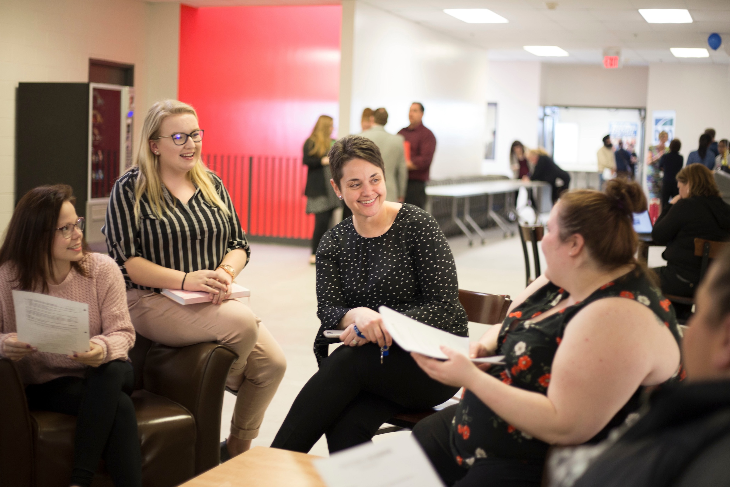 A group of people are sitting down in a chairs in a circle and speaking to one another. There are other groups of people speaking to each other in the same room behind the close-up of the group.