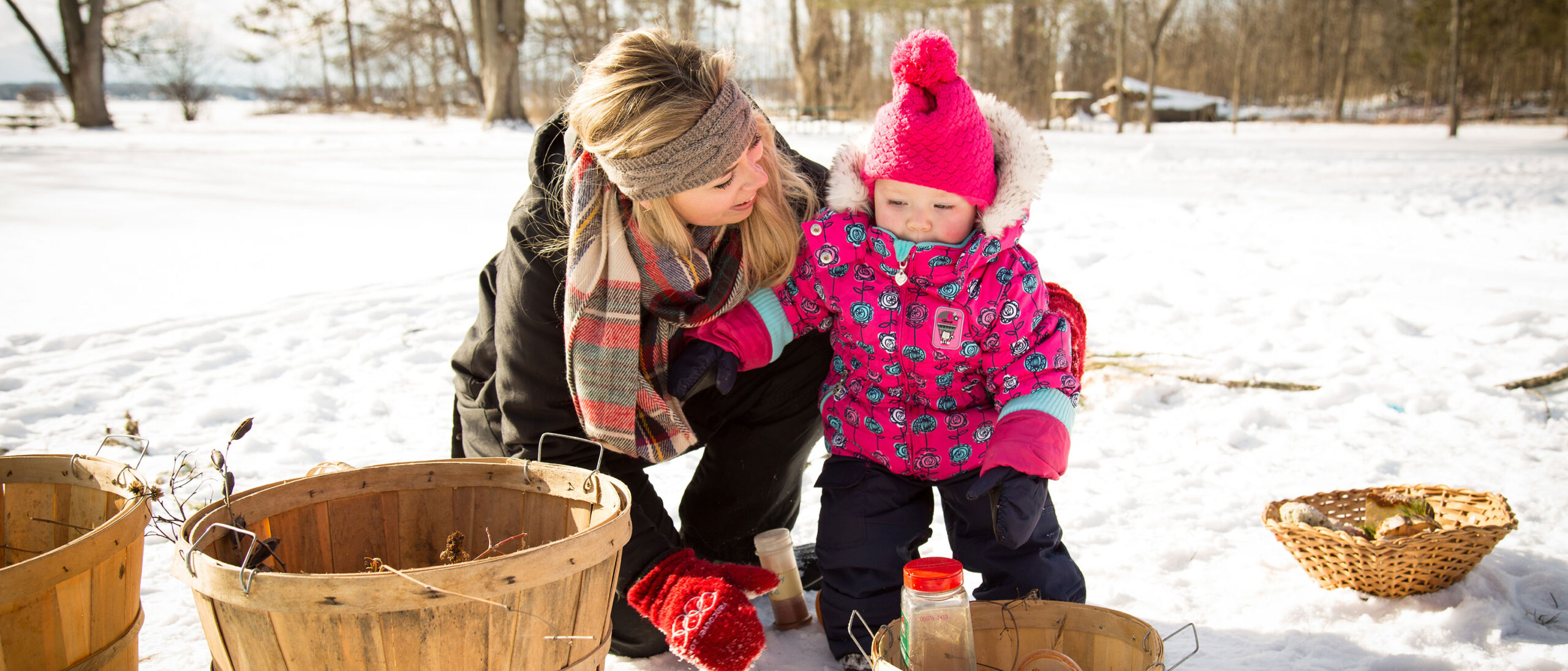 An early childhood education student is smiling and working with a baby outside in the winter. They are both wearing winter gear, surrounded by baskets and snow.