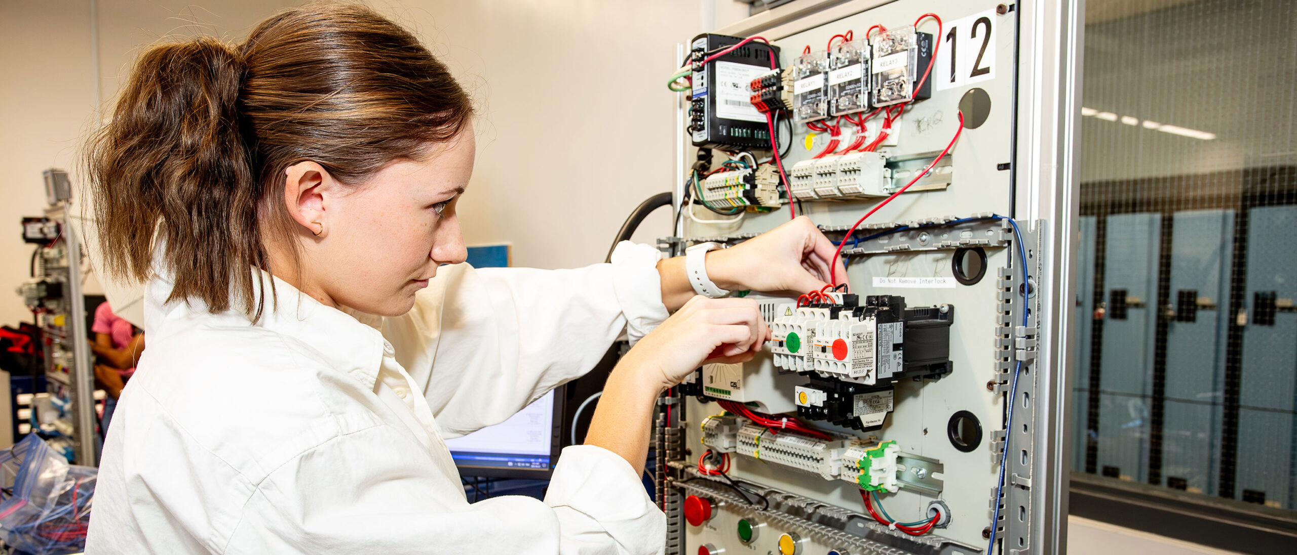 An electrical engineering student is working with wires and circuits inside a classroom setting. The student is wearing a white lab coat with their hair tied up in a ponytail.