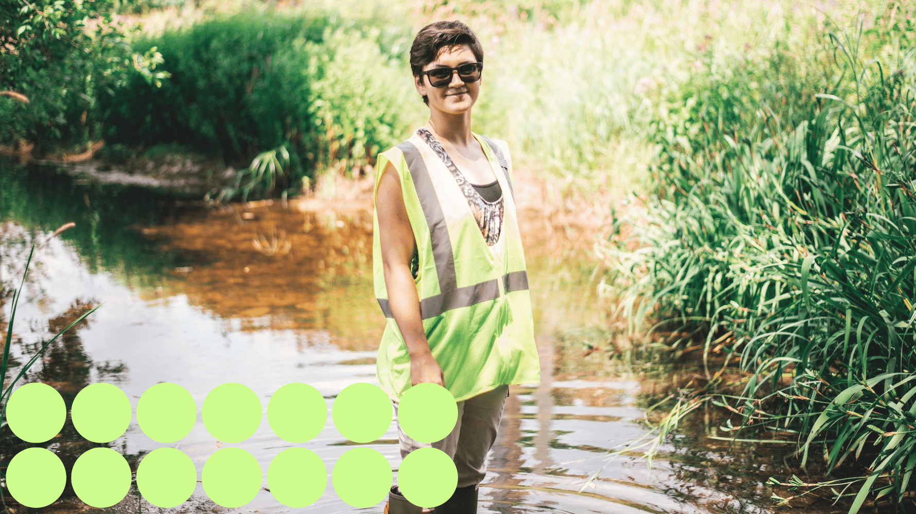 A student in a high visibility vest with sunglasses, stands in a stream with rubber boots on, smiling at the camera. Green graphic circles are in the lower lefthand corner of the image.