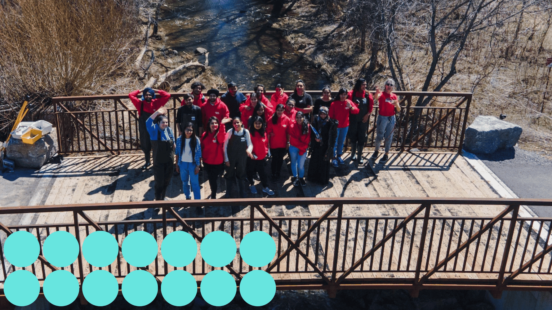 Loyalist students, mostly in red shirts, are standing on a wooden bridge over a stream, looking up at the camera. Blue graphic circles are in the lower lefthand corner of the image.