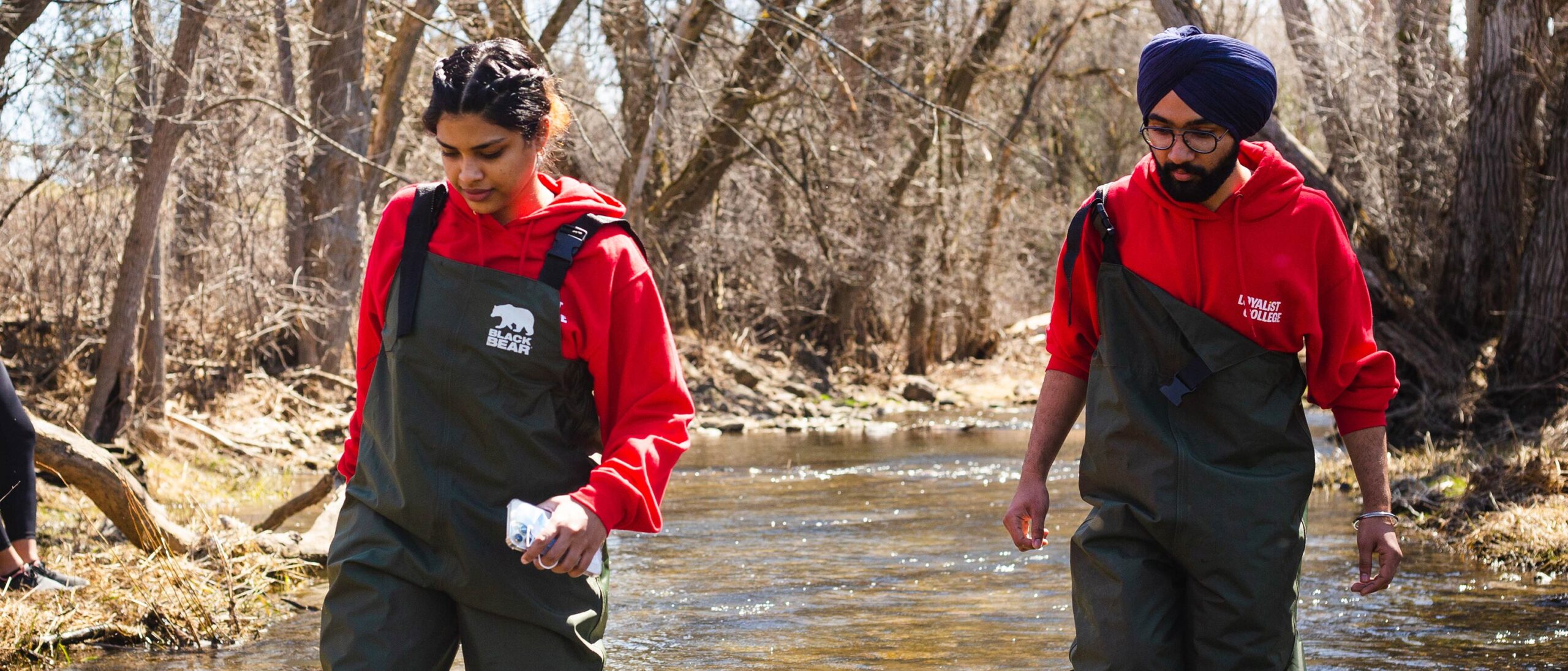 Two students walking through a shallow river, both wearing bright red hoodies and black waterproof coveralls. They carefully navigate the rocky riverbed, with the water reaching just below their knees. The vibrant red of their hoodies contrasts with the natural surroundings, adding a pop of colour to the serene outdoor setting.