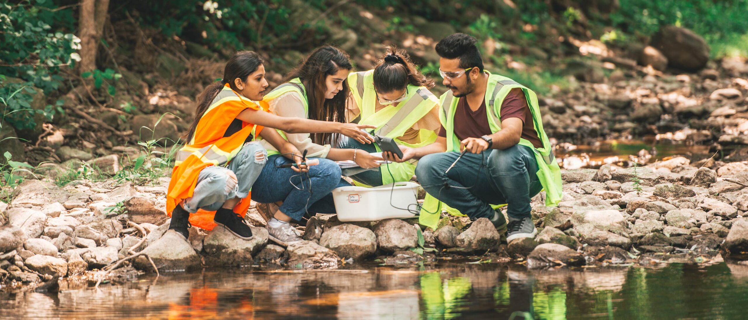 A group of four students in the environmental technology program are working on getting samples in an outdoor setting. The students are wearing neon construction vests, kneeled down on rocks, overlooking a river.