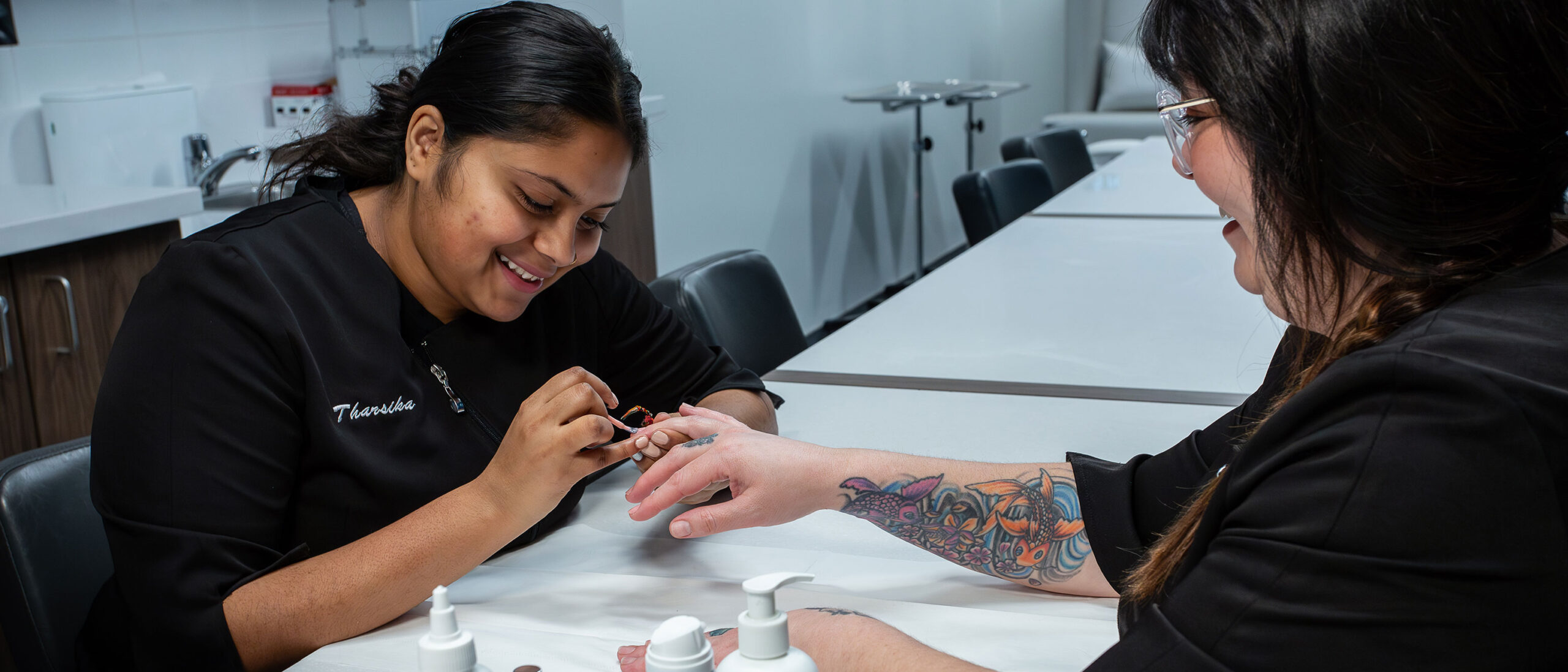 An esthetics and spa management student is painting a mock client's nails. The student and mock client are both wearing black t-shirts and smiling.