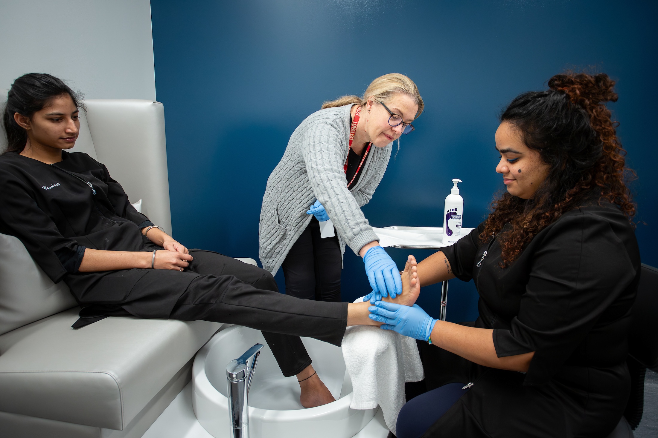 Two Loyalist esthetics students and an esthetics professors are pictured. One of the students is sitting on a bench with her feet in a small sink on the ground. The other student is being guided by the professor while holding one of the girls feet, going over techniques.