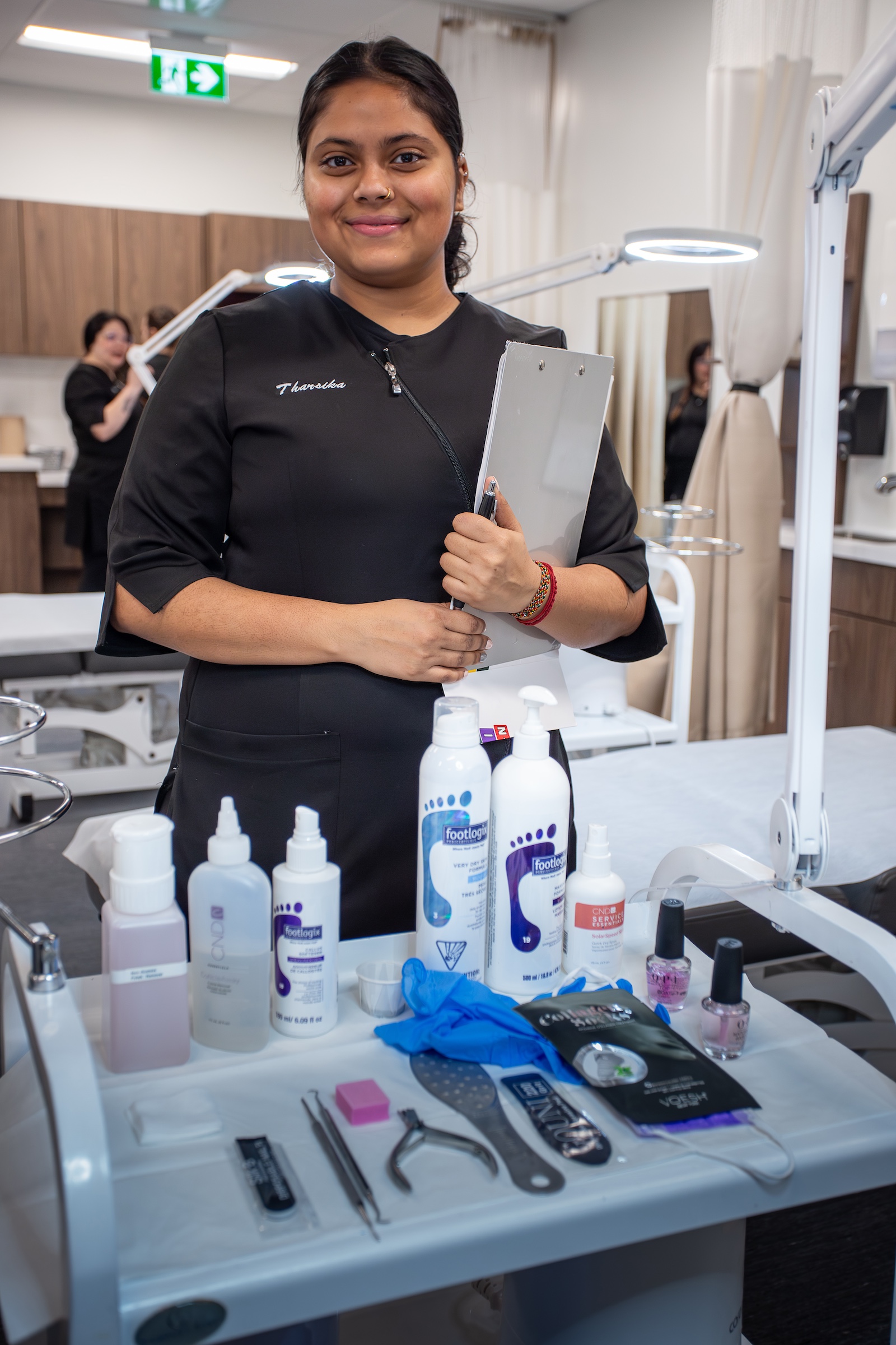 Loyalist esthetics student in a black uniform holding a clipboard in front of a tray of tools and lotions for foot care.