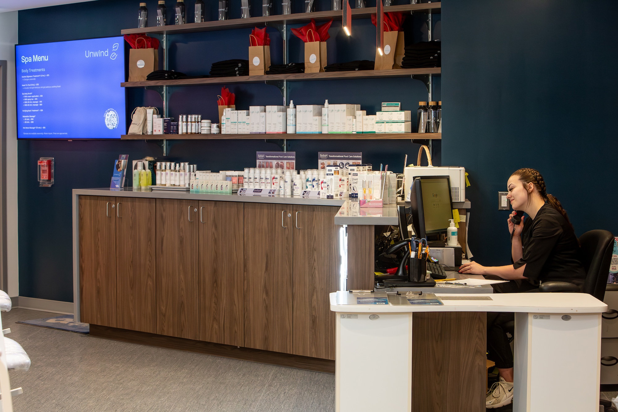 A Loyalist esthetics student sitting behind the desk of the Unwind Spa at Loyalist. There are shelves with various products on them on the wall and a TV screen showing a Spa Menu.