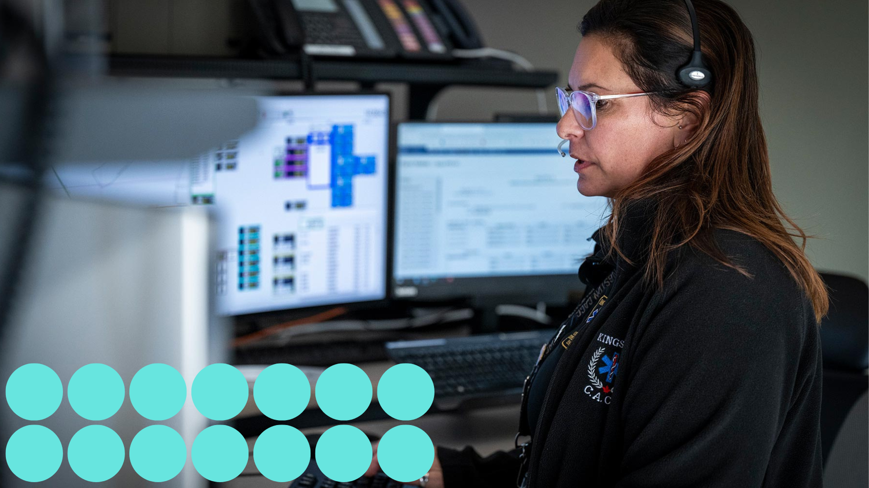 A female 911 dispatcher, wearing a headset and glasses, is focused on multiple computer monitors at a dispatch centre.