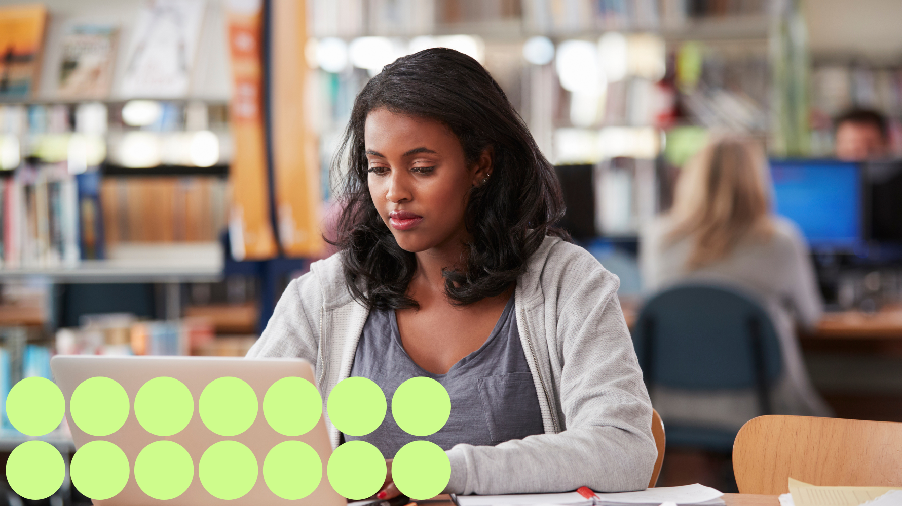 A student is sitting down in the library working on their laptop. There are other students in the background using computers. On the back left of the image are bookshelves. Fourteen light green graphic circles are along the bottom left of the image.