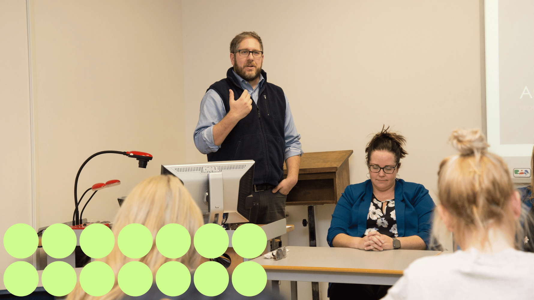 A professor stands at the front of the room speaking before a desk containing a computer, with a person seated next to him and students listening in front of him and taking notes. Green graphic circles are in the lower lefthand corner of the image.