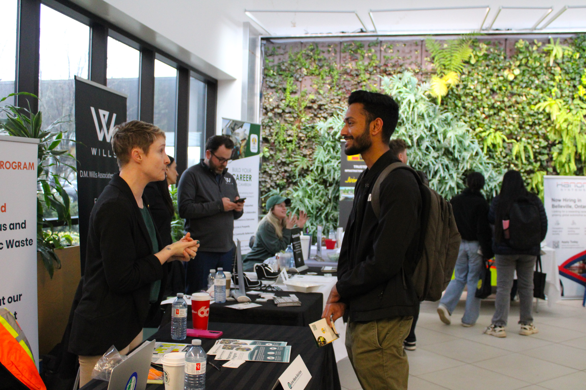 A student engages in a conversation with a representative at a booth during a campus career fair. The representative is standing behind a table covered with informational brochures and water bottles. Other students and representatives are visible in the background, and the event is held in a well-lit space with a large green wall of plants.