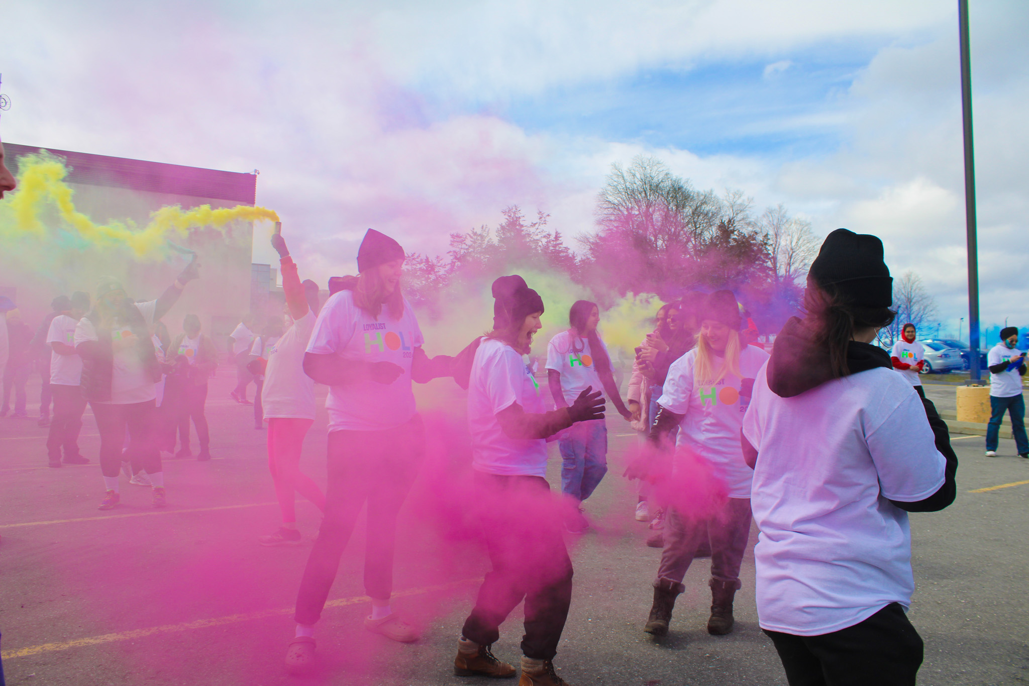 A group of students is joyfully celebrating during a Holi event, surrounded by vibrant clouds of pink, yellow, and purple powder. The students, dressed in white T-shirts, are fully immersed in the festivities, laughing and dancing as they throw coloured powder into the air.