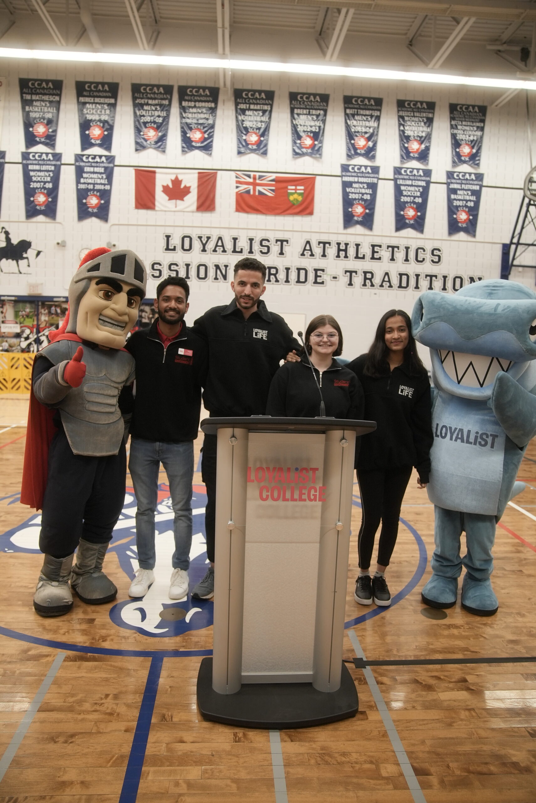 Group of students standing in a gymnasium alongside two mascots—one dressed as a knight and the other as a shark—posing for a photo in front of a podium with the Loyalist College logo.