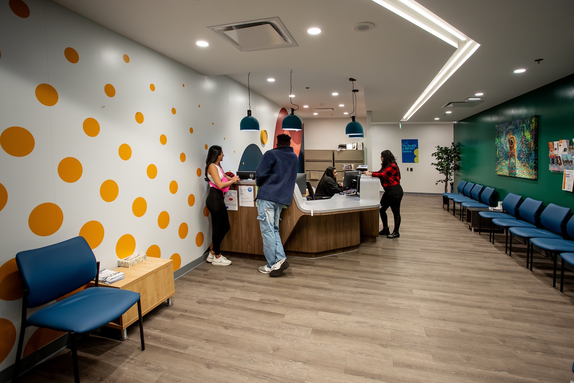 Three students stand at the reception desk in the health centre, where another person works behind the desk. Empty chairs line the long room for students to wait in.