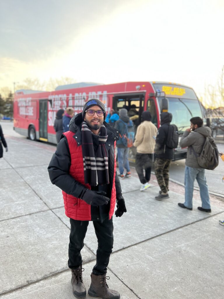 A student in a winter jacket and hat smiles at the camera in front of a line of students boarding a large red bus.