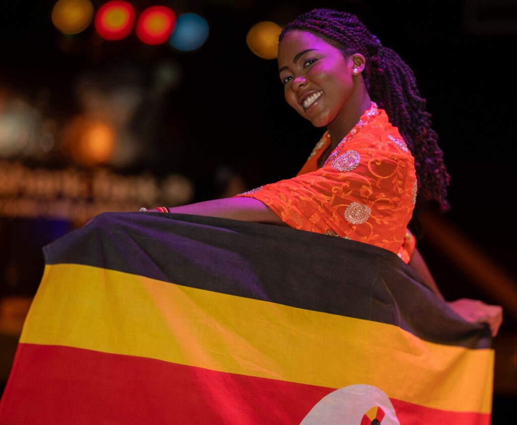 Person in a vibrant orange outfit smiling warmly, holding a large Uganda flag. The background is softly blurred with colorful lights, creating a festive atmosphere, at a cultural event.