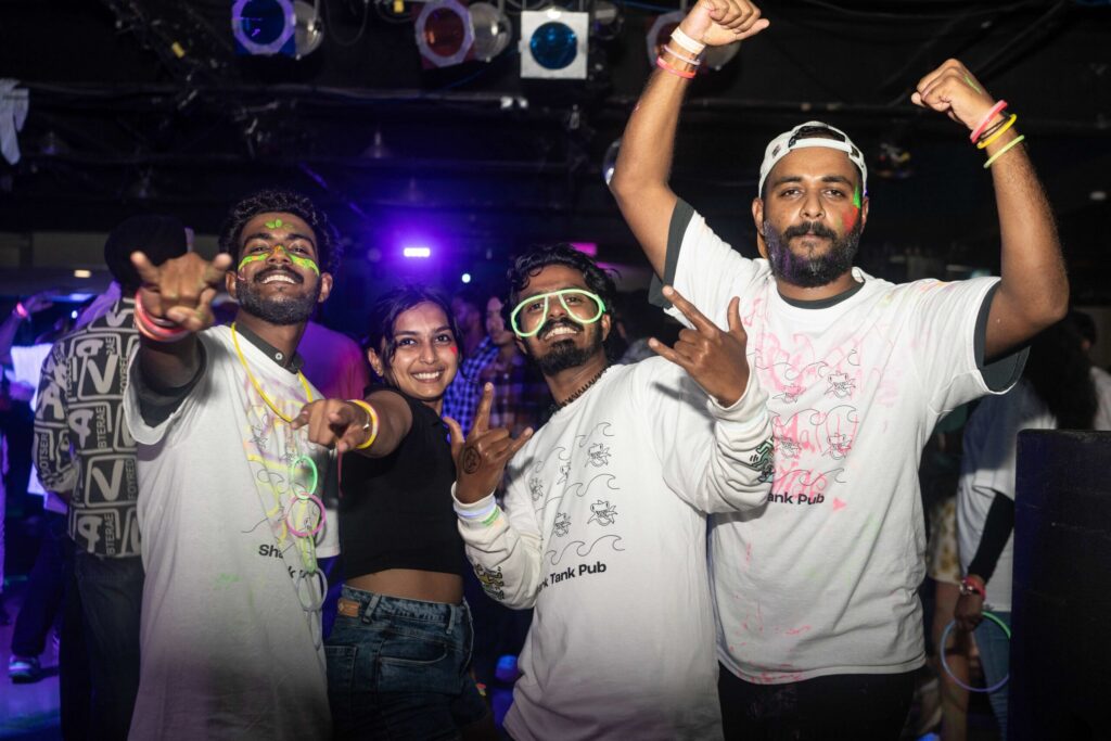 A group of friends enjoying a lively night out, wearing neon accessories and face paint. They are smiling, pointing, and posing energetically in a vibrant, club-like atmosphere with colourful lights in the background.