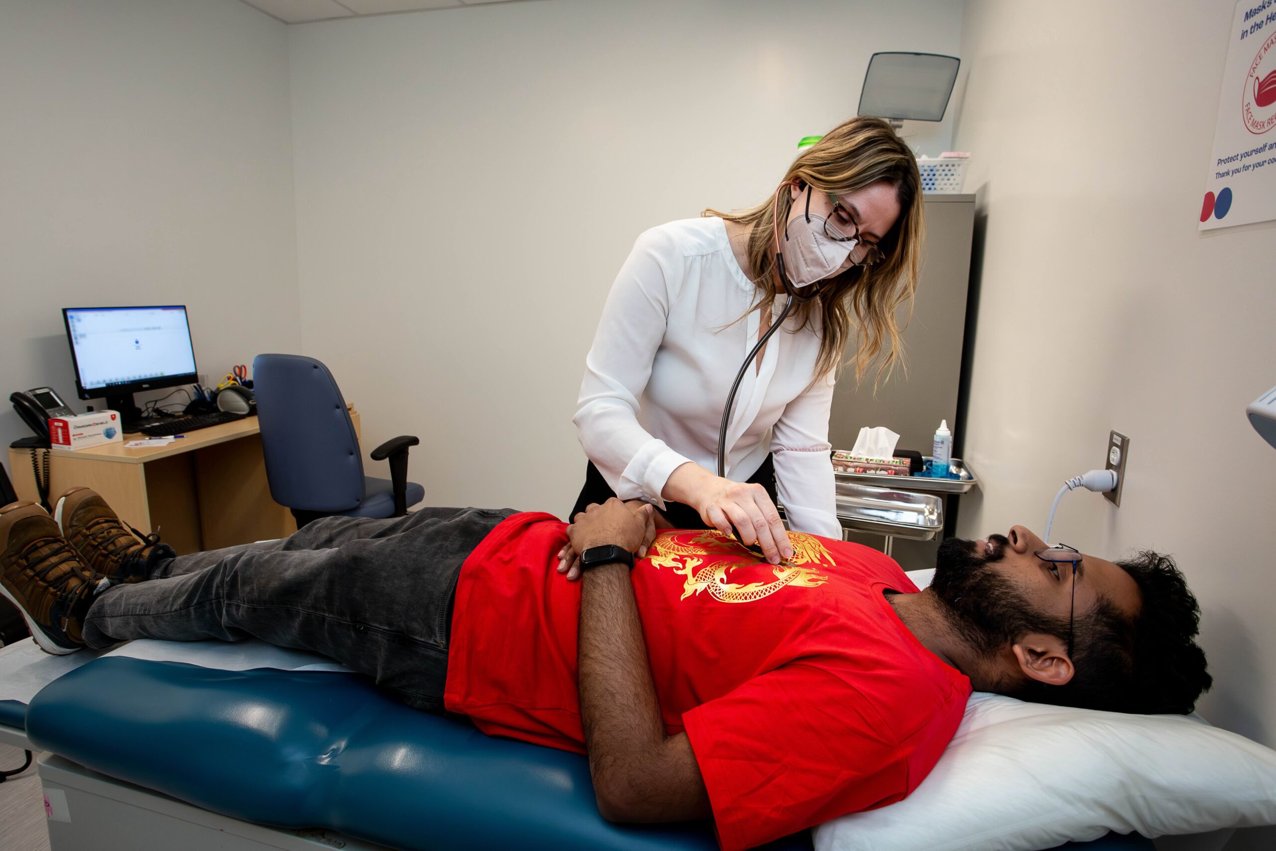 The image shows a healthcare professional, wearing a white blouse and a protective face mask, using a stethoscope to examine a patient lying on an examination table. The patient, who is dressed in a red shirt with a gold design, is relaxed and looking upwards while the healthcare provider listens to their chest. The setting appears to be a medical examination room, with a computer on a desk, medical supplies on a cart, and a wall poster promoting health safety visible in the background.