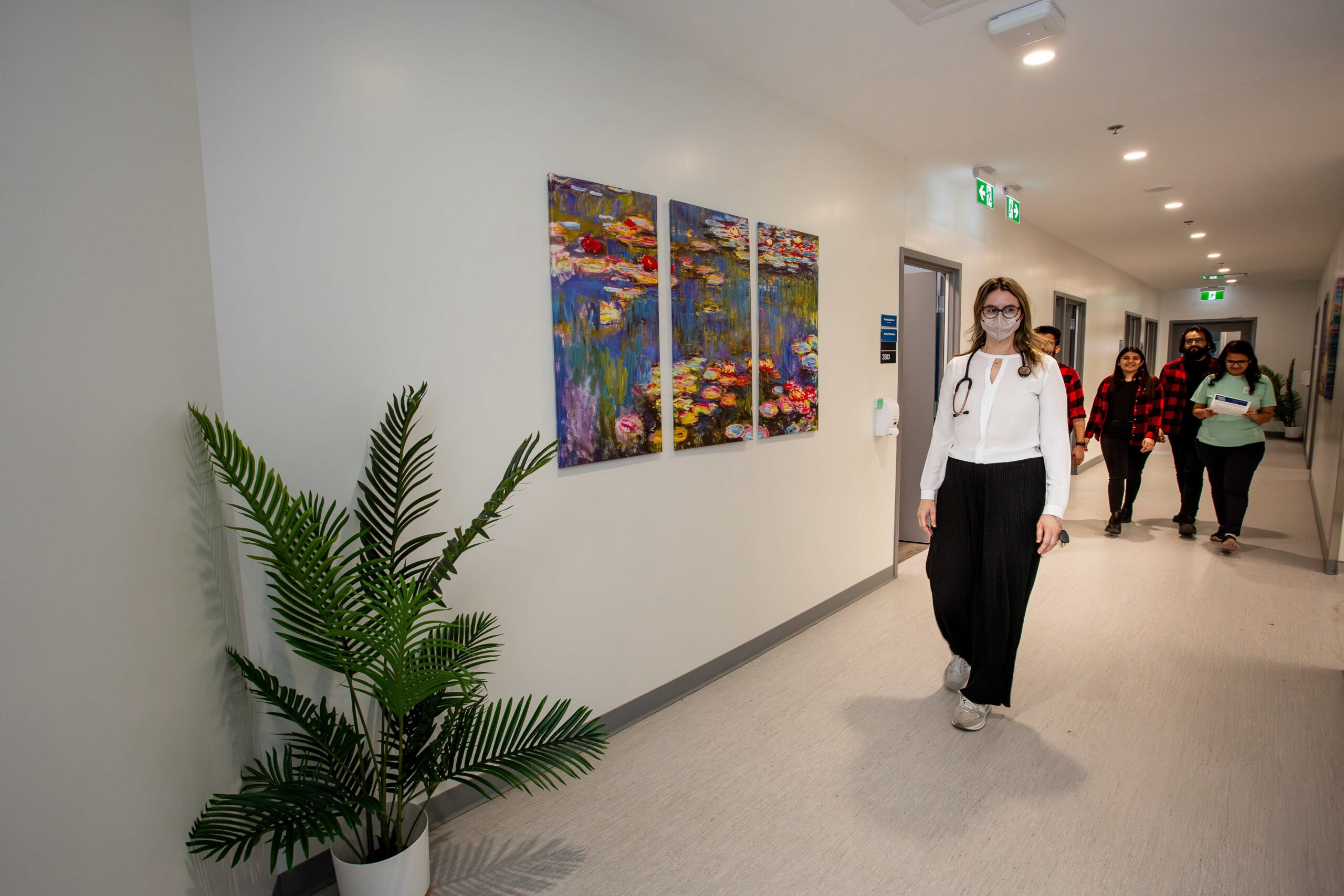 The image depicts a hallway in a healthcare or wellness facility. A healthcare professional, dressed in a white blouse, black pants, and wearing a stethoscope around her neck, is walking toward the camera with a confident and approachable demeanor. She is wearing a face mask. Behind her, three people are walking down the hallway, engaged in conversation and carrying papers. The hallway is clean and well-lit, with a large green plant placed near the wall, and colorful artwork depicting water lilies hanging on the wall, adding a calming and welcoming atmosphere to the space.