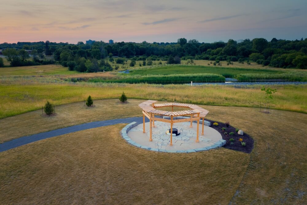 A beautiful wood circle pergola stands on a stone and concrete platform with grass around it and a path leading to it. The sun is setting in the background.