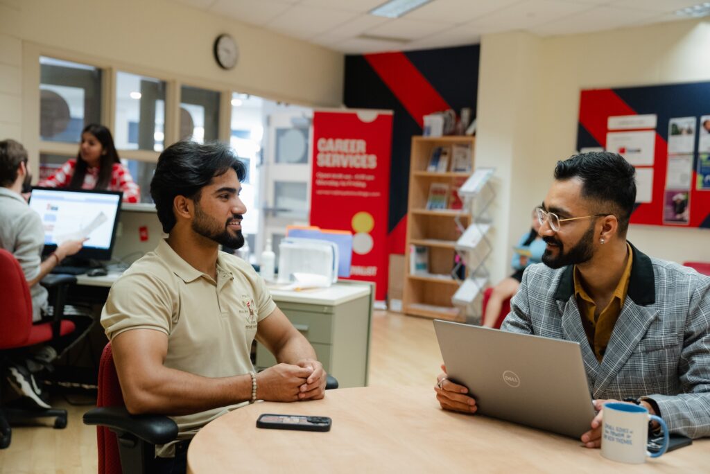 The main focus of the image show two people sitting down across from each other at the table talking to each other. In the background there is another duo speaking to one another. By the door of the room, there is a pop-up banner that says, "career services."